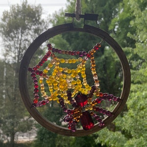 Puede incluir: Decoración de telaraña de cuentas. La telaraña está hecha con cuentas amarillas, naranjas y rojas, y está suspendida dentro de un aro de bordado de madera. Una araña de cuentas con cuerpo rojo y patas moradas se encuentra en el centro. Se adjunta un lazo de hilo natural para colgar.