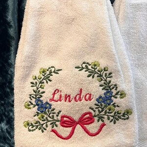 May include: White hand towel with floral and butterfly print at the top. The towel features an embroidered wreath with the name "Linda" in pink, a red bow, and green and blue floral accents.