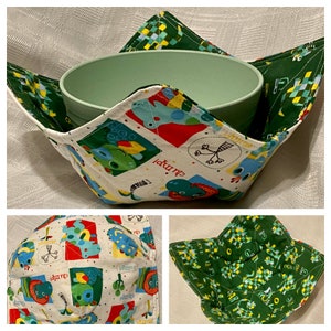 Puede incluir: Un calientaplatos de tela verde y blanco con un patrón de dinosaurio. El calientaplatos está doblado para mostrar el interior, que es verde con un patrón a cuadros. El calientaplatos está diseñado para sostener un tazón.