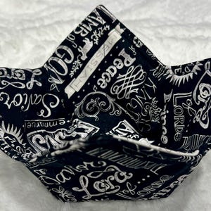 May include: Black and white fabric bowl with a variety of religious words and phrases printed on it. The words include "Lord", "Savior", "Peace", and "Love".