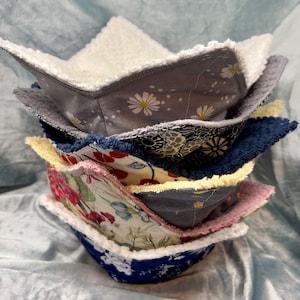 Może przedstawiać: Stos sześciu miseczek wykonanych z tkaniny w różnych wzorach kwiatowych. Miski są wykonane z miękkiej, pikowanej tkaniny z białym polarem. Miski są włożone jedna w drugą, największa na dole, a najmniejsza na górze.