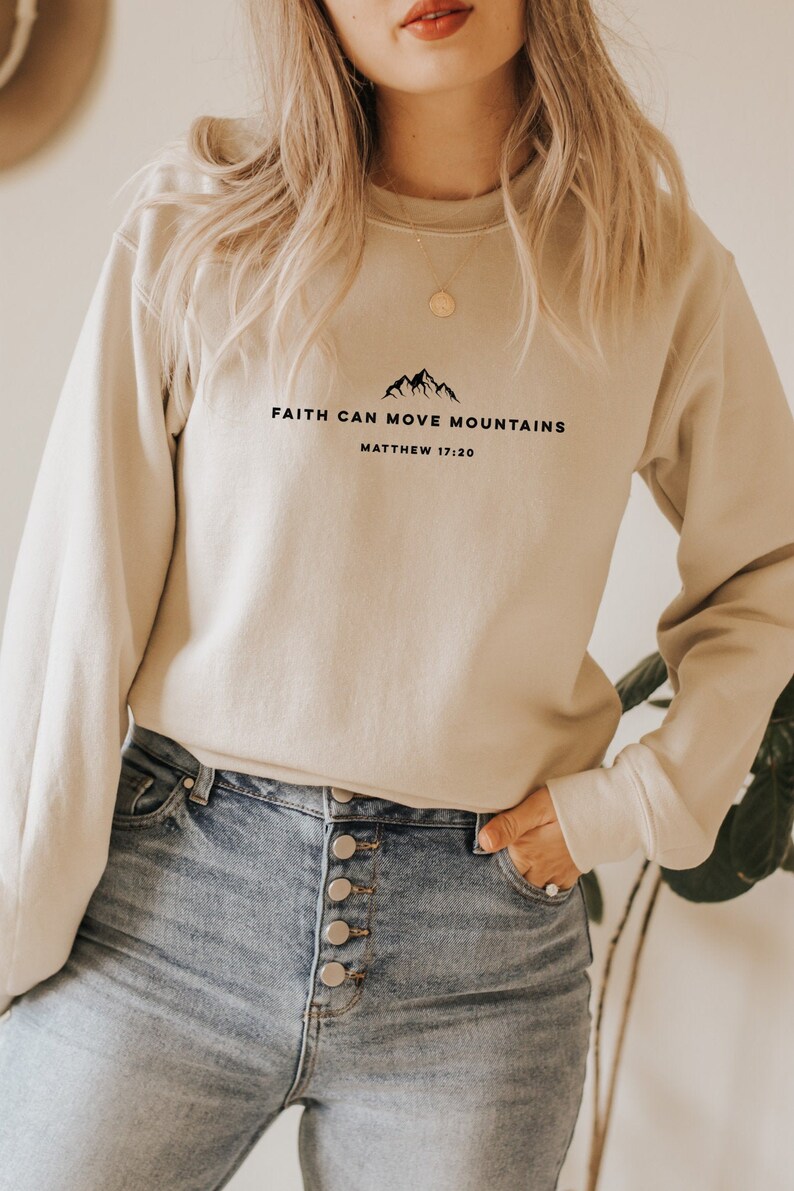 May include: A beige sweatshirt with the text "FAITH CAN MOVE MOUNTAINS" and "MATTHEW 17:20" printed on the front. The sweatshirt is being worn by a person with long blonde hair.