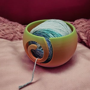 May include: A green and orange yarn bowl with a ball of blue and green yarn inside. The yarn is threaded through a hole in the bowl.