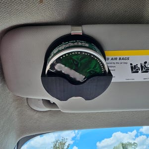 May include: A black plastic car visor clip holding a can of Long Cut Wintergreen. The can is green and silver with text and a graphic. The clip is attached to a gray car visor.