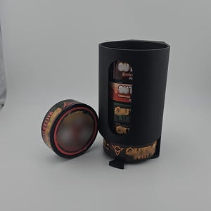 May include: A black container with a gold-colored base and a slot revealing several small, cylindrical containers. The containers have labels like "Orange" and "Win". A separate, similar container with "Watermelon" text is also present.