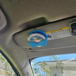 May include: A blue car visor clip holding a small, round mirror. The clip is attached to a car visor, with a warning label visible. The mirror reflects a blurred image. The clip is made of plastic.