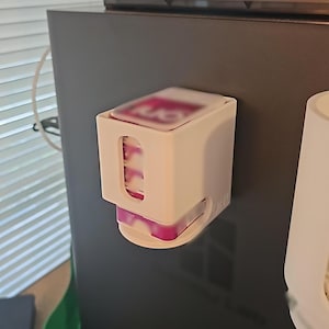 May include: A white plastic card holder with a square design, mounted on a dark gray surface. The holder contains pink and white cards, with the word "UNO" visible on the top card. The holder has a curved bottom.