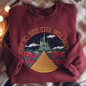 May include: Burgundy sweatshirt with an embroidered circular design. The design features a yellow brick road leading to a green castle under a starry night sky, surrounded by red poppies. The text "OFF TO SEE THE WIZARD" is embroidered around the circle.