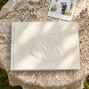 May include: A rectangular cream-colored fabric photo album featuring an embroidered design of two swans facing each other. The text "The Smith" and "EST. 2026" is embroidered in a script font above the swans on the front cover of the album.