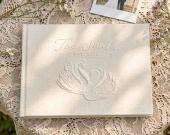 May include: A rectangular cream-colored fabric photo album featuring an embroidered design of two swans facing each other. The text "The Smith" and "EST. 2026" is embroidered in a script font above the swans on the front cover of the album.