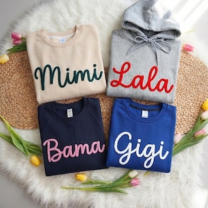 May include: Four sweatshirts and a hoodie in neutral colors, each embroidered with a name. The sweatshirts are beige, navy blue, and royal blue. The hoodie is gray. The names are embroidered in green, red, pink, and white. The items are arranged on a white fur-like surface.