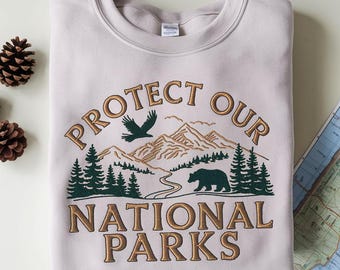 Può includere: Felpa beige chiaro con la scritta ricamata "PROTECT OUR NATIONAL PARKS". Il design presenta una catena montuosa, alberi, un orso e un uccello in volo, nei toni del verde e del marrone.