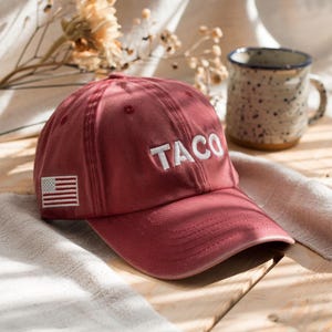 May include: A maroon baseball cap with the word "TACO" embroidered in white. The cap features a small American flag patch on the side. The hat is made of a soft material and is suitable for casual wear.