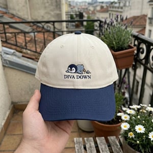May include: A cream-colored baseball cap with a navy blue brim. The cap features an embroidered penguin design with the text "DIVA DOWN" in black. The cap has a curved brim and a small navy blue button on top.