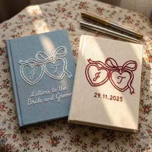 May include: Two hardcover books with fabric covers. The blue book features white embroidered hearts with the letters J and T and the text Letters to the Bride and Groom. The cream book features red embroidered hearts with J and T and 29.11.2025.