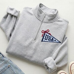 May include: A gray quarter-zip sweatshirt with a blue and white gingham pennant embroidered with "USA" and a red bow. The sweatshirt is folded with the sleeves cuffed. A pair of jeans and sneakers are visible in the image.