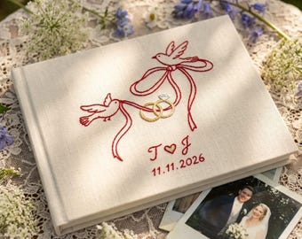 May include: A cream-colored fabric-covered photo album featuring red embroidered birds holding two gold rings with a diamond, the initials T and J, and the date 11.11.2026. The cover design includes flowing red ribbons connecting the birds and rings.