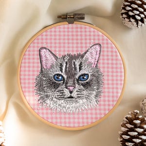 May include: An embroidered cat portrait in a wooden hoop. The cat has gray fur, blue eyes, and pink ears and nose. The background is a pink and white gingham pattern.