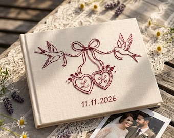 May include: A rectangular cream-colored fabric-covered photo album featuring dark red embroidery of two birds holding a ribbon tied to two heart-shaped lockets with the letters L and H inside, and the date 11.11.2026 embroidered below.