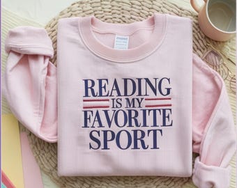 May include: A light pink sweatshirt with the text "READING IS MY FAVORITE SPORT" in navy blue and red. The sweatshirt is folded on a woven mat, with a pink mug and glasses in the background.