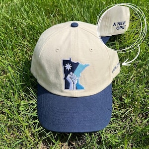 May include: A beige and navy blue baseball cap with a curved brim. The cap features an embroidered design of the Minnesota state outline, a star, and a raised fist. The text "A NEW OPER" is embroidered on the side.