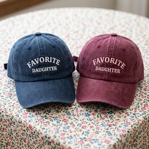 May include: Two baseball caps, one navy blue and one burgundy, are shown. Each cap features the words "FAVORITE DAUGHTER" embroidered in white. The caps are placed on a floral patterned surface.