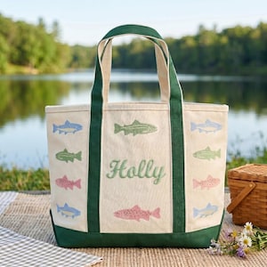 May include: Cream-colored tote bag with green trim and handles. The bag features embroidered fish in various colors and the name "Holly" in green script. The bag is sitting on a picnic blanket near a lake.