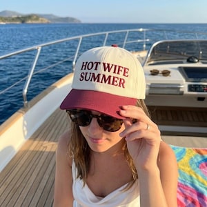 May include: A cream-colored baseball cap with a maroon brim and the text "HOT WIFE SUMMER" in maroon lettering. The cap is worn on a boat with a blue ocean in the background.