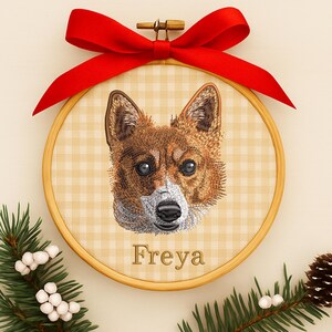 May include: Embroidered dog portrait in a wooden hoop, featuring a brown and white dog with the name "Freya" below. The hoop is adorned with a red ribbon bow at the top and surrounded by greenery and berries.