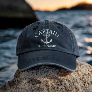 May include: A navy blue baseball cap with the word "CAPTAIN" embroidered in white above an anchor design. The anchor has a gold-colored rope. Below the anchor, the text "YOUR NAME" is embroidered in white. The cap is sitting on a rock.