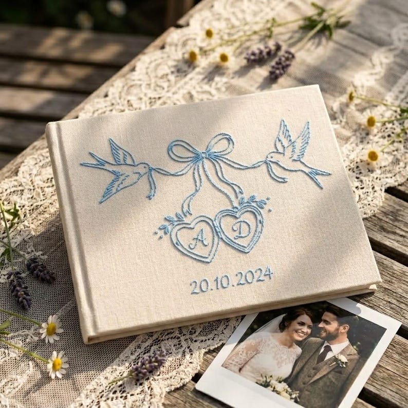 May include: A rectangular beige fabric-covered photo album featuring light blue embroidery of two birds holding a ribbon tied to two hearts. The hearts contain the letters A and D, with the date 20.10.2024 embroidered below in blue thread.