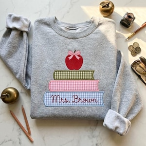 May include: A gray sweatshirt with a stack of books and a red apple design. The books are in green, pink, and blue gingham patterns, with the name "Mrs. Brown" embroidered on the bottom book. A pink bow sits atop the apple.