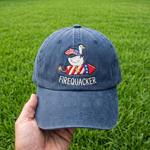 May include: A navy blue baseball cap with an embroidered design of a white duck riding a red, white, and blue rocket. The duck wears an American flag as a cape. The word "FIREQUACKER" is embroidered below the rocket.