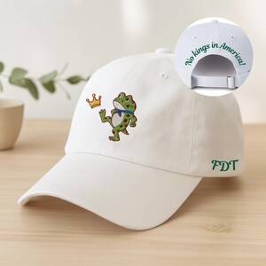 May include: White baseball cap with an embroidered frog wearing a blue scarf and a crown. The text "No kings in America!" is on the back, and "FDT" is embroidered on the side. A fun, casual accessory.