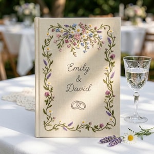 May include: A linen-covered wedding guest book with embroidered floral details. The cover features the names "Emily & David" and intertwined rings. The floral embroidery includes daisies, lavender, and other flowers in shades of pink, blue, and white.