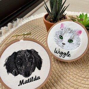 May include: Two embroidered hoops featuring pet portraits. One depicts a black and white dog with the name "Matilda" below. The other shows a gray cat with pink ears and the name "Wiggle". Both are set in wooden hoops.