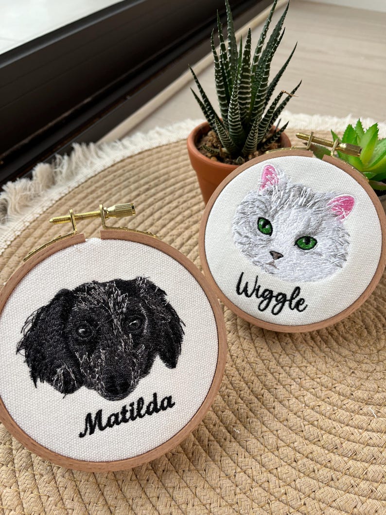 May include: Two embroidered hoops featuring pet portraits. One depicts a black and white dog with the name "Matilda" below. The other shows a gray cat with pink ears and the name "Wiggle". Both are set in wooden hoops.