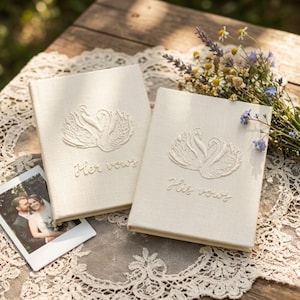 May include: Two cream-colored fabric-covered vow books featuring an embroidered design of two swans facing each other. One book is embroidered with the text Her vows and the other with His vows. A small photograph of a couple is placed nearby.