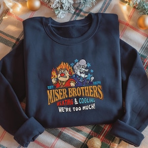 May include: Navy blue sweatshirt featuring an embroidered design of the Miser Brothers from the 1974 animated film. The design includes the text "Miser Brothers Heating & Cooling" and "We're Too Much!"