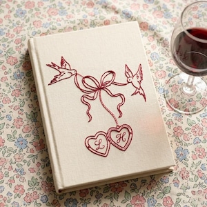 May include: A cream-colored hardcover book featuring red embroidery of two birds holding a ribbon tied to two heart-shaped lockets. The left locket contains the letter L and the right locket contains the letter H, both stitched in red thread.