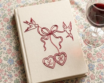 May include: A cream-colored hardcover book featuring red embroidery of two birds holding a ribbon tied to two heart-shaped lockets. The left locket contains the letter L and the right locket contains the letter H, both stitched in red thread.