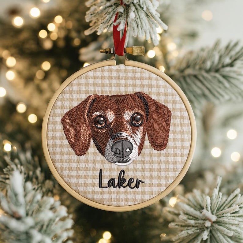 May include: Embroidered dog portrait ornament in a wooden hoop. The dog has brown fur, a black nose, and the name "Laker" embroidered below. The background is a tan and white gingham pattern. A red ribbon is attached for hanging.