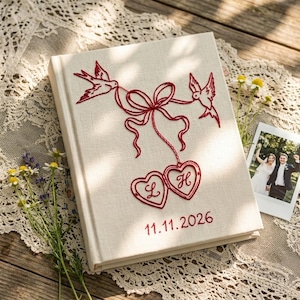 May include: A cream-colored fabric-covered photo album featuring red embroidered details. The design includes two birds holding a ribbon tied into a bow, which connects to two heart-shaped frames containing the letters L and H, and the date 11.11.2026.