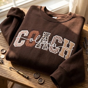 May include: A dark brown crewneck sweatshirt featuring the word COACH in large, block-style letters. Each letter has a unique pattern, including floral, polka dot, leopard print, plaid, and tan dots. A white cursive script overlays the letters.