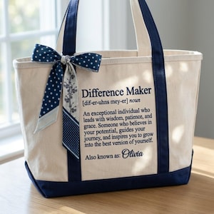 May include: A cream-colored canvas tote bag with navy blue trim and handles. A navy blue and white polka dot ribbon is tied to the handle. The bag features the printed text: Difference Maker [dif-er-uhns mey-er] noun, followed by a definition and the name Olivia.