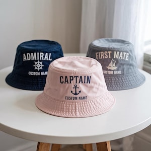 May include: Three bucket hats in navy, pink, and gray, each with nautical-themed text. The navy hat says "ADMIRAL" with a ship's wheel, the pink hat says "CAPTAIN" with an anchor, and the gray hat says "FIRST MATE" with a sailboat. All hats have "CUSTOM NAME" below the text.