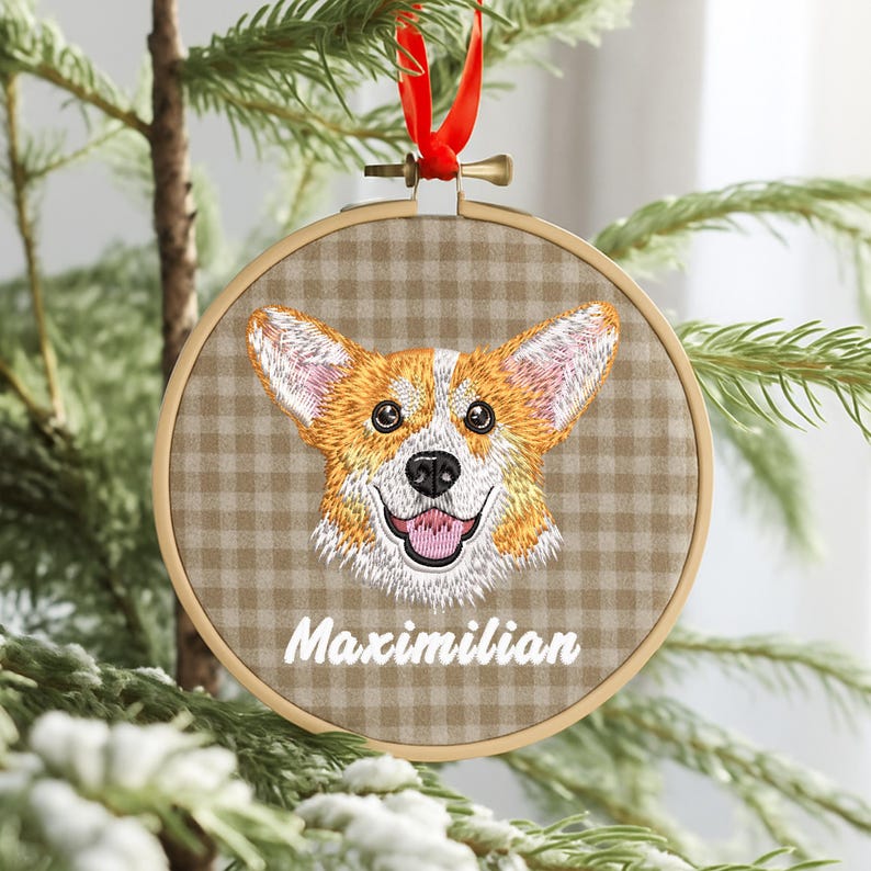 May include: A round, embroidered ornament featuring a smiling corgi dog face with orange, white, and black details. The ornament is set in a wooden hoop with a brown and tan plaid background. The name "Maximilian" is embroidered below the dog's face. A red ribbon is attached for hanging.