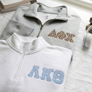 May include: A grey quarter-zip sweatshirt with embroidered Greek letters in a leopard print pattern and a white quarter-zip sweatshirt with embroidered Greek letters in a light blue and white polka dot pattern, both featuring high collars.