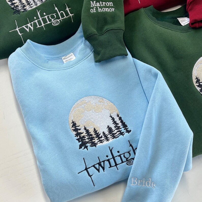 May include: Light blue sweatshirt with an embroidered design of a full moon and pine trees. The word "twilight" is embroidered in black script across the front. The sleeve says "Bride". Other sweatshirts in green and burgundy are visible.