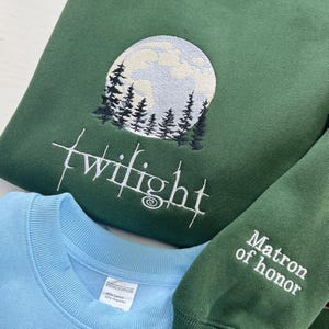 May include: Green sweatshirt with a white embroidered design of a full moon, trees, and the word "Twilight." A light blue sweatshirt is visible below. The sleeve of a green sweatshirt reads "Matron of honor."